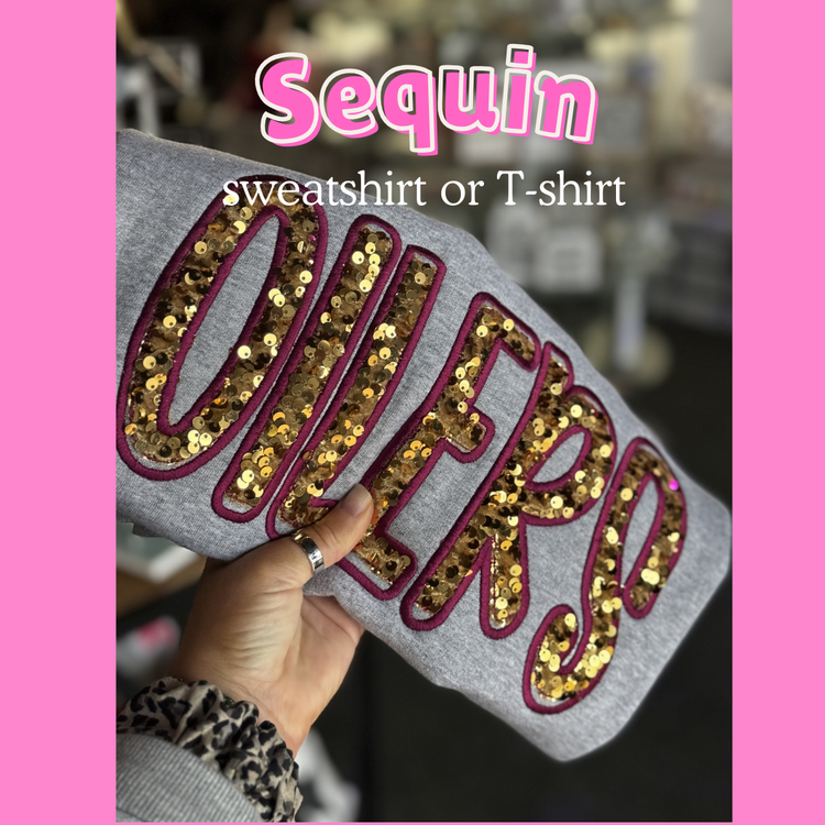 Female Hand holding folded gray shirt that says Oilers with gold sequins and a maroon thread outline. 